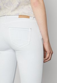 Bílé denimové džíny s přiléhavým střihem, které mají zadní kapsu, decentní švy a světlou hnědou koženou nášivku na pase.