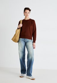 Sudadera marrón con escote en V, combinada con unos vaqueros de pierna ancha de un azul claro y desgastado. El modelo lleva un bolso tote acolchado en color tostado. Zapatillas de deporte plateadas completan el look.