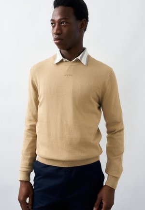 Muž v béžovom roláku s logom „HUGO“ cez bielu košeľu s golierom, v tmavomodrých nohaviciach, pozerajúci sa do strany.