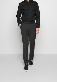 Maglietta nera a maniche lunghe con chiusura a bottoni, abbinata a pantaloni grigi su misura e sneakers nere con suola bianca. Design semplice, taglio aderente.