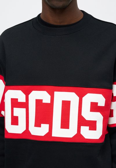 Μαύρο φούτερ με κόκκινη λωρίδα που φέρει μεγάλες λευκές μπλοκ γραφές "GCDS." Το ύφασμα έχει υφή με ραμμένα μανίκια.