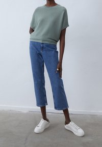 Osoba ubrana w jasnozielony prążkowany top, niebieskie obcisłe dżinsy, białe trampki i złote pierścienie na palcu, stojąca na betonowej podłodze.