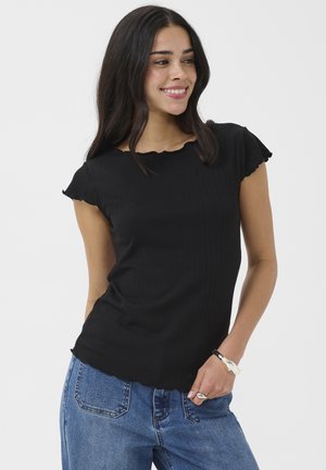 Šypsanti moteris su ilgais tamsiais plaukais, dėvinti juodą ribotą trumpomis rankovėmis palaidinę ir mėlynas džinsus, stovinti su viena ranka kišenėje.