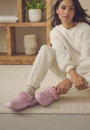 Frau sitzt auf einem beigen Teppich, trägt cremefarbene Freizeitkleidung und zieht rosa, mit Fell gefütterte Clogs an, im Hintergrund ein Holzregal und eine Pflanze.