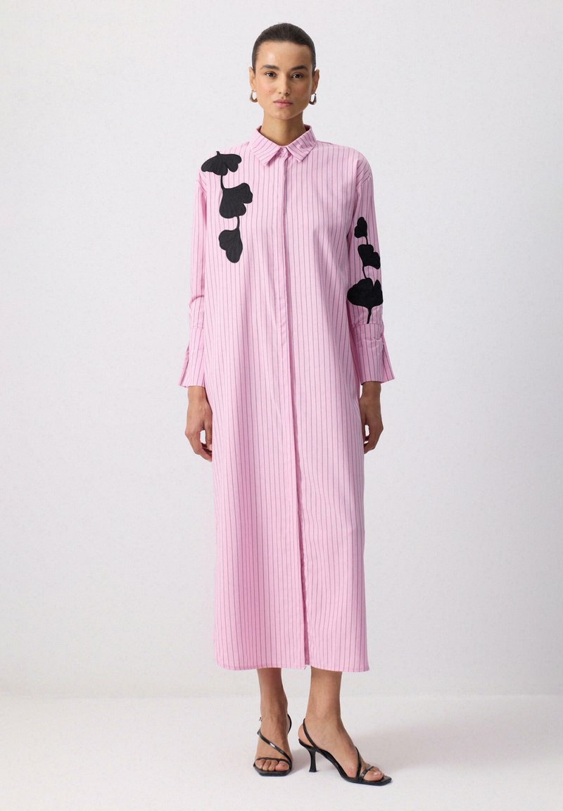 Femeie purtând o rochie lungă roz cu dungi subțiri și modele negre de frunze pe mâneci și umeri, asortată cu pantofi cu barete negre.