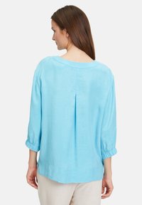 Light blue, loose-fitting blouse with three-quarter sleeves, a wide neckline, and a center back pleat. Fabric has a soft, smooth texture.