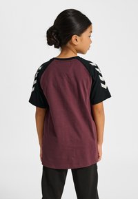 Vinröd t-shirt med svarta raglanärmar, som har vita chevronränder på axlarna. Mjukt tyg, ledig passform och rund halsringning.