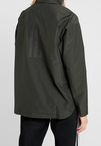 Mørkegrøn, letvægtsjakke med høj krave, der har en mat finish og tre lodrette striber trykt på ryggen.