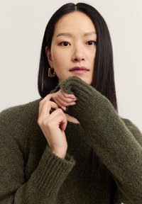 Grüner gestrickter Pullover mit einem gerippten Rundhalsausschnitt und langen Ärmeln, der eine strukturierte Oberfläche aufweist. Das Model hält die Ärmel an den Handgelenken.