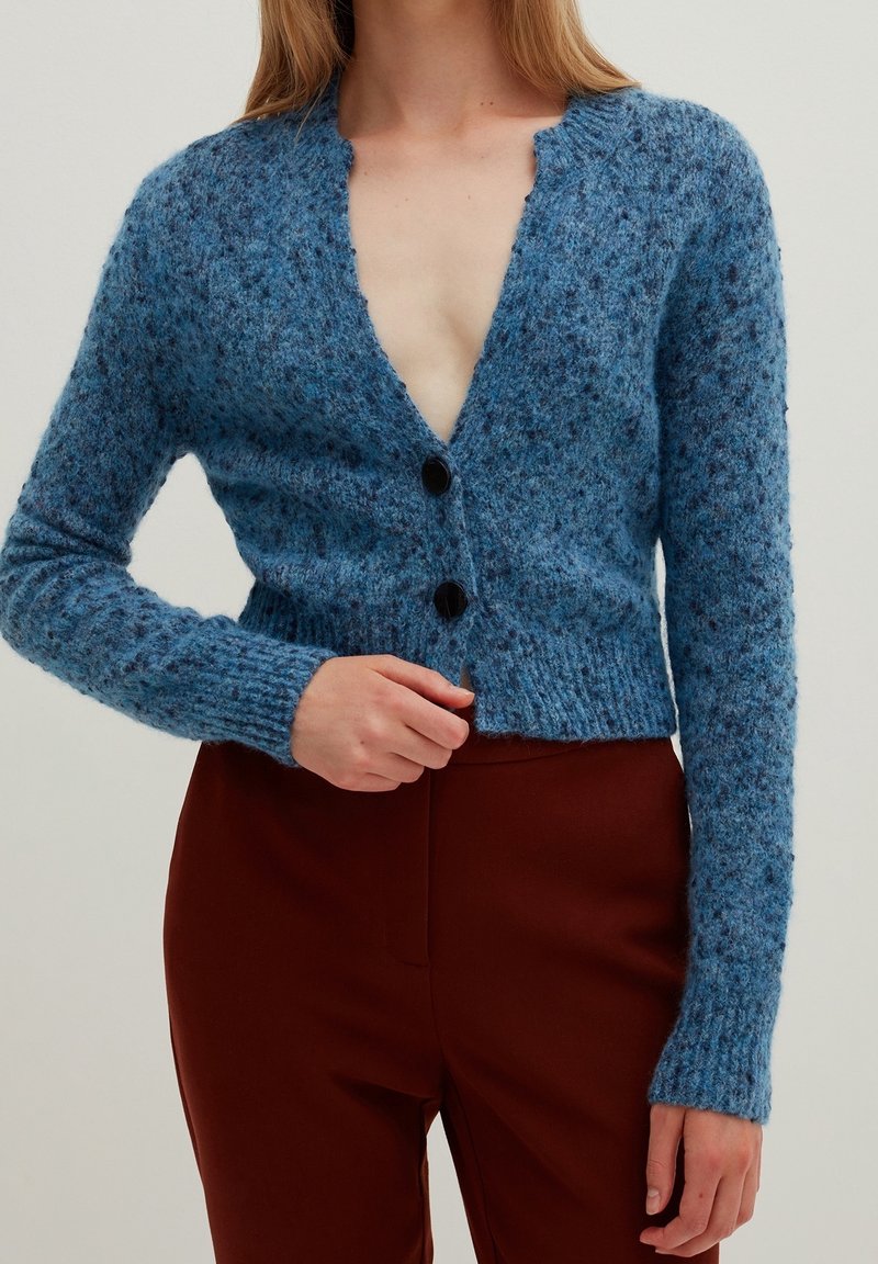 Kobieta ubrana w teksturowany niebieski krótki kardigan z czarnymi guzikami na brązowych spodniakach z wysokim stanem, ręka dostosowująca przód kardiganu.