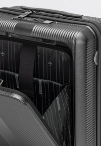 Valigia rigida nera con texture a righe e doppia cerniera. L'interno presenta una fodera nera con motivi di aerei bianchi e del marchio.