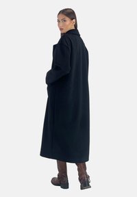 Abrigo largo de lana negra con cuello y bolsillos laterales. La modelo lleva botas de cuero marrón hasta la rodilla con detalles de hebilla. Tela suave y entallada.