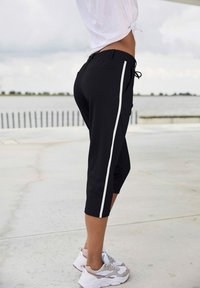 Zwarte capri-broek met witte zijkant, elastische tailleband en trekkoord. Gepaard met wit en grijze sportschoenen. Soepele stof textuur.