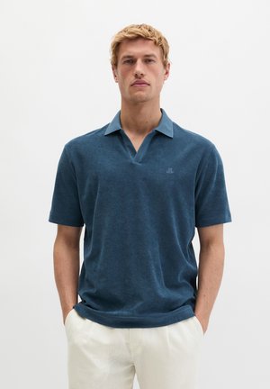 Blondyn w niebieskiej koszulce polo z krótkim rękawem i białych spodniach, stojący z rękami w kieszeniach na jednolitym tle.