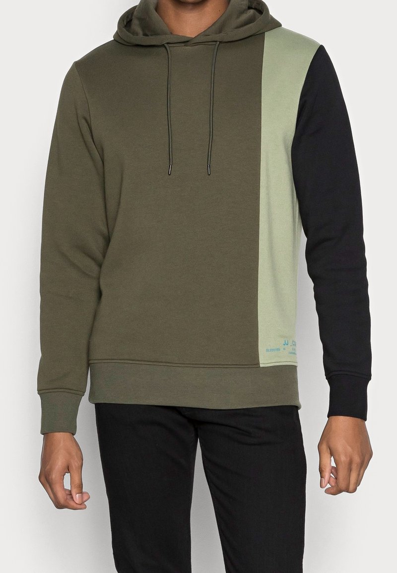 Homme portant un sweat à capuche bicolore avec un devant vert olive, un panneau latéral vert clair, une manche droite noire et un pantalon noir.