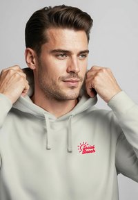 Hellegrauer Kapuzenpullover mit verstellbarem Kordelzug und rotem "Social Beach"-Logo auf der Brust. Weicher Stoff, lässiges Design.