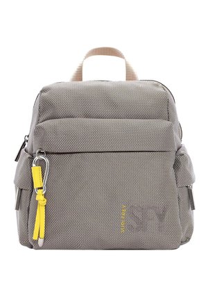 Personne portant un jean bleu, un t-shirt beige, des baskets blanches et un sac à dos gris avec une fermeture éclair jaune, debout devant un fond uni.