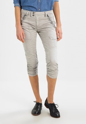 Vrouw draagt lichtbeige cropped cargobroek met ritssluitingen, gecombineerd met zwarte veterschoenen en een blauwe denim blouse.