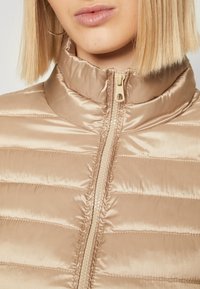 Helle beigefarbene gesteppte Jacke mit hohem Kragen und Kunststoffreißverschluss. Besitzt eine glatte, glänzende Textur und horizontale Nähte.