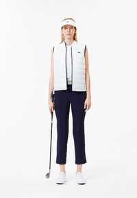 Witte gewatteerde vest over een wit shirt, gecombineerd met navy slim-fit broek. Model houdt een golfclub vast; draagt een witte visord en witte schoenen.