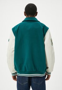 Groene en cream varsityjack met een wollen lichaam, leren mouwen, gestreepte manchetten en een geborduurd nummer op de mouw.