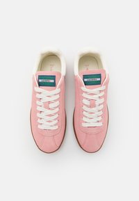 Zapatillas de ante rosa con cordones blancos, que cuentan con una etiqueta del logo de Lacoste en verde y azul en la lengüeta y una suela de goma marrón.