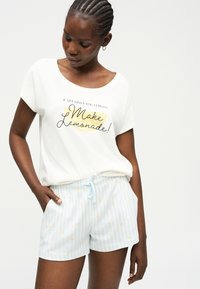 Γυναίκα με πλεγμένα μαλλιά που φοράει λευκό μπλουζάκι με την επιγραφή "Αν η ζωή σου δίνει λεμόνια, φτιάξε λεμονάδα!" και ανοιχτά μπλε ριγέ σορτς με κίτρινες κουκκίδες.