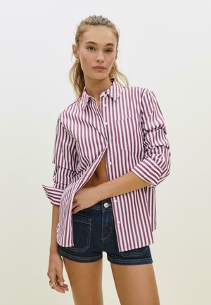 Camicia a righe bordeaux e bianca, con maniche arrotolate e taschino sul petto; abbinata a pantaloni corti in denim scuro.