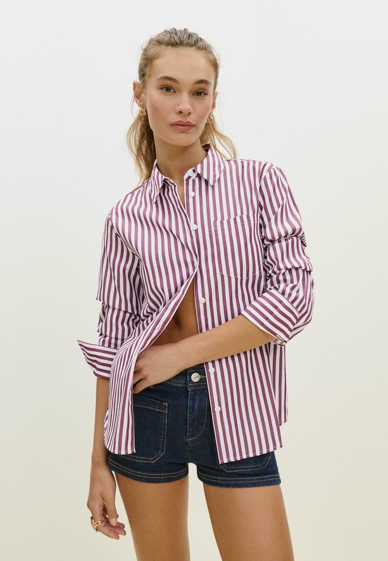 Camicia a righe bordeaux e bianca, con maniche arrotolate e taschino sul petto; abbinata a pantaloni corti in denim scuro.