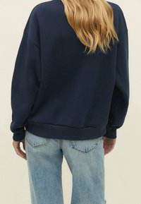 Persona con lunghi capelli biondi indossa un felpa blu navy e jeans in denim azzurri chiari, in piedi contro uno sfondo chiaro e uniforme.