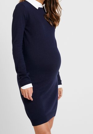 Kobieta w ciąży w granatowej sukience z długim rękawem z białym kołnierzykiem i mankietami, stojąca na tle jasnego, jednolitego tła.