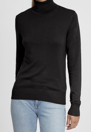 Femme portant un pull à col roulé noir associé à un jean en denim bleu clair, debout devant un fond gris clair uni.