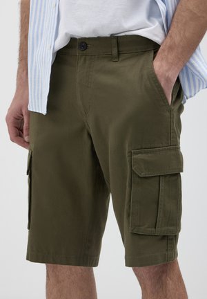 Homme portant un short cargo vert olive avec des poches latérales, un T-shirt blanc et une chemise rayée bleu clair, avec une main dans la poche.