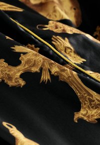 Čierna látka s komplikovanými zlatými vzormi a detailnými motívmi. Dopĺňa to tenký žltý zips. Hladká textúra, bohatý dizajn.