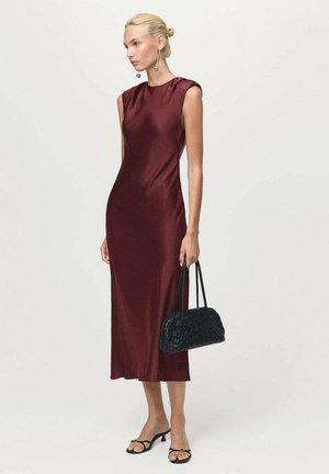 Frau in einem ärmellosen burgunderfarbenen Midikleid, die eine schwarze gewebte Handtasche hält, schwarze Absatzsandalen trägt und silberne Ohrhänger trägt.