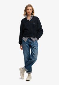 Granatowy sweter w warkocz z dekoltem w serek, nałożony na paskowaną koszulę z guzikami, zestawiony z luźnymi niebieskimi dżinsami i kremowymi Sneakersami w stylu wysokim.