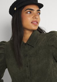 Olivengrønn corduroy-skjorte med knapper foran, krage og puffede ermer. Modellen har på seg en svart hatt med gullakenter.