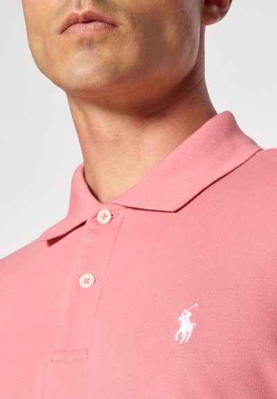 Camisa polo rosa feita de tecido texturizado, com colarinho, dois botões na pala e um logótipo bordado em branco no peito.