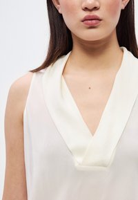 Κρεμ αμάνικη μπλούζα με γιακά σε σχήμα V, με λεία, απαλή υφή και μινιμαλιστικό σχέδιο.