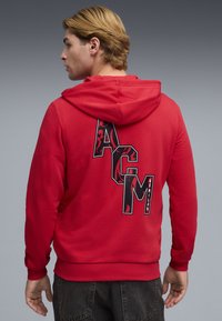 Κόκκινο φούτερ με κουκούλα και κορδόνι, με μεγάλο γραφικό σχέδιο "ACM" σε μαύρο και σχέδιο στο πίσω μέρος. Μαλακό ύφασμα και ριμπ μανσέτες.