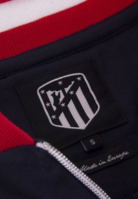 Marineblauwe zip-up jas interieur met rood en wit gestreepte ribbel; heeft een geborduurd zwart-wit schildlogo en maatlabel.