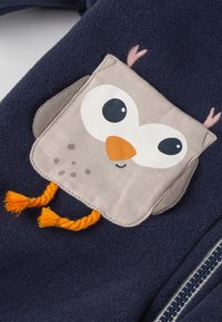 Dunkelblaues Fleece-Jackett mit einem cremefarbenen Eulen-Patch, das einen orangefarbenen Schnabel und Füße sowie pinkfarbene Ohrbüschel hat; seitlicher Reißverschluss.