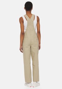 Salopette en coton beige avec bretelles réglables, grandes poches avant et jambes amples. Associée à un débardeur blanc et des baskets.