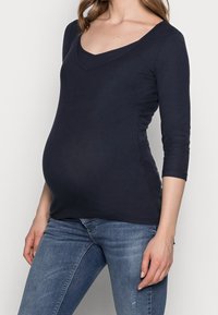 Marineblaues Longsleeve-Oberteil mit V-Ausschnitt, geripptem Muster und figurbetontem Schnitt, speziell für Umstandsmode entworfen, kombiniert mit blauen Jeans.