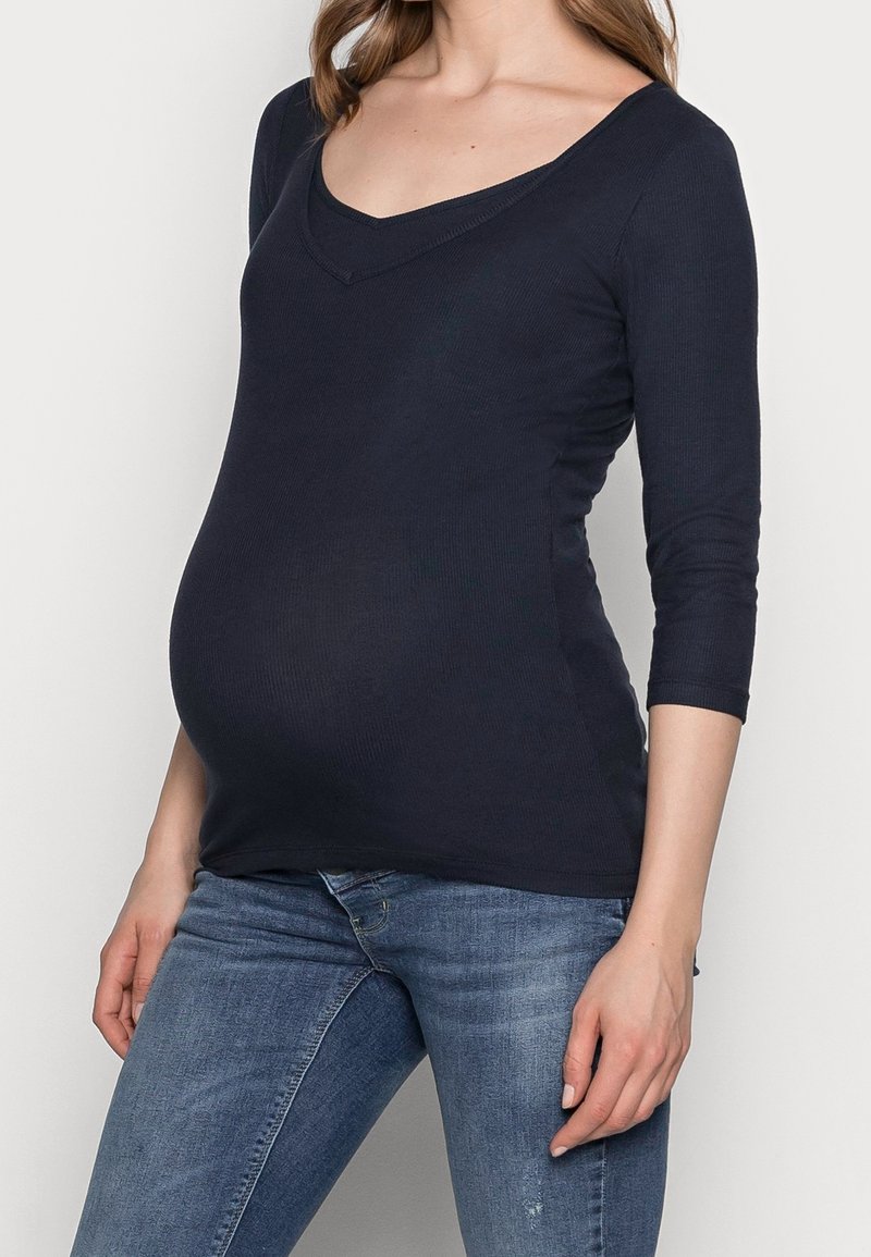 Marineblaues Longsleeve-Oberteil mit V-Ausschnitt, geripptem Muster und figurbetontem Schnitt, speziell für Umstandsmode entworfen, kombiniert mit blauen Jeans.