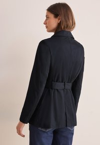 Blazer blu scuro con colletto sartoriale, dettagli con cintura in vita, maniche lunghe e un tessuto leggermente testurizzato. Design semplice e classico.