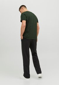 Camiseta de algodón verde oscuro de manga corta, combinada con pantalones negros. La persona está de pie con las manos en los bolsillos, vista desde atrás.