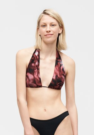 Blondynka ubrana w czerwono-czarny wzorzysty stanik bikini z wiązaniem na szyi oraz czarne, jednolite majtki bikini na jednolitym tle.