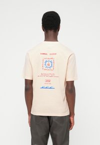 Άνδρας με κοντά σγουρά μαλλιά, φοράει μπεζ μπλουζάκι με κόκκινο και μπλε γραφικό κείμενο και κύματα, στέκεται με την πλάτη προς το μέρος μας σε απλό φόντο.