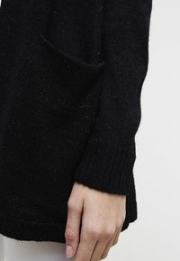 Černý pletený svetr s žebrovanou texturou, volným střihem a boční kapsou viditelnou na levé straně, dlouhými rukávy a rovnou spodní linií.